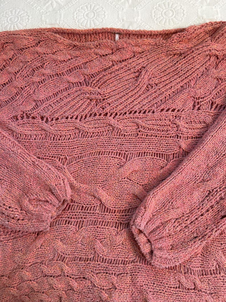 Puede incluir: Un su&eacute;ter de color rosa empolvado con un dise&ntilde;o de punto texturizado. El su&eacute;ter presenta un cuello barco y mangas largas con un patr&oacute;n de punto trenzado. La prenda de punto tiene un aspecto suave y ligeramente descolorido.