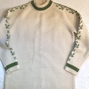 May include: Cream-colored sweater with green accents. The sweater features a high collar and cuffs with green stripes. Green cross-like patterns adorn the shoulders and sleeves. The sweater has a ribbed hem and cuffs.