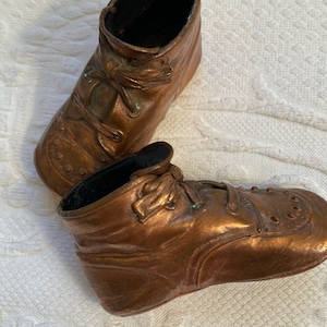 Peut inclure: Une paire de chaussures de bébé en cuivre antique. Les chaussures sont détaillées avec des lacets et ont une patine usée et vieillie.