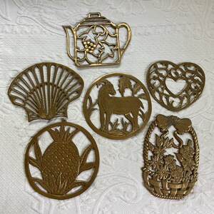 May include: Six brass trivets with intricate designs. The trivets feature a teapot, a seashell, a goat, a pineapple, a heart, and a basket of flowers.