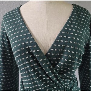May include: A dark green wrap top with long sleeves and a V-neck. The fabric features a pattern of small, evenly spaced white dashes. The top is displayed on a mannequin, highlighting the design and fit of the garment.