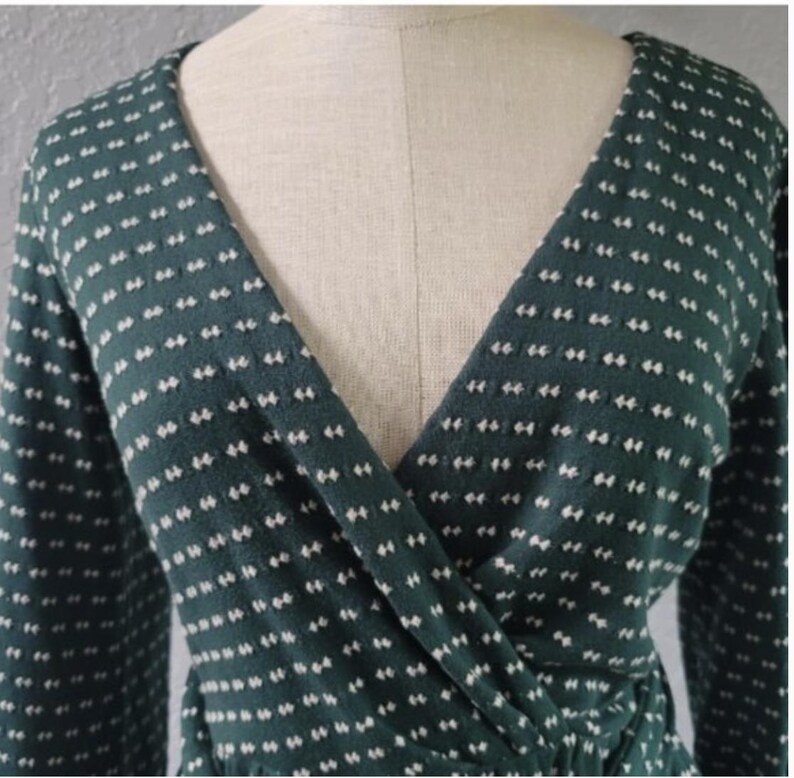 May include: A dark green wrap top with long sleeves and a V-neck. The fabric features a pattern of small, evenly spaced white dashes. The top is displayed on a mannequin, highlighting the design and fit of the garment.