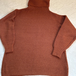 May include: A rust-colored turtleneck sweater with a ribbed knit texture. The sweater has long sleeves with ribbed cuffs and a wide, folded turtleneck collar. The garment is made of a thick, warm material, perfect for cooler weather.