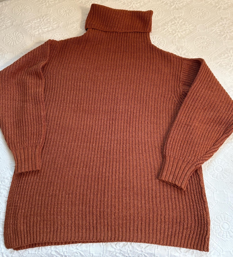 May include: A rust-colored turtleneck sweater with a ribbed knit texture. The sweater has long sleeves with ribbed cuffs and a wide, folded turtleneck collar. The garment is made of a thick, warm material, perfect for cooler weather.