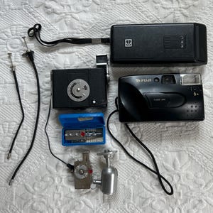 Op de afbeelding: Een verzameling vintage camera's, waaronder een zwarte en zilveren Fuji filmcamera met flitser, een zwarte en zilveren camera met een wijzerplaat, een zwarte en zilveren cameratas en een zilveren camera-accessoire met een rode knop.