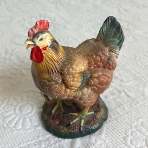 May include: A brown and white ceramic figurine of a hen with a red comb and green tail feathers. The hen is standing on a brown base with green accents.