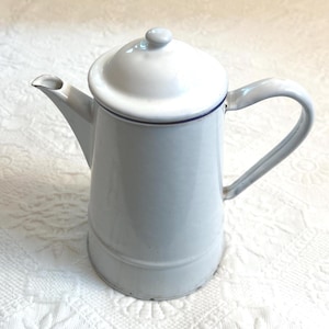 Op de afbeelding: Een witte emaille koffiepot met een deksel en een gebogen handvat. De pot heeft een tuit en een donkerblauwe rand rond het deksel. De pot is lichtblauw.