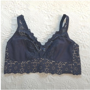 May include: A navy blue bra with lace detailing. The bra features a scalloped lace trim along the neckline and bottom edge. The cups and straps are also adorned with delicate lace patterns. The bra is designed with adjustable straps.