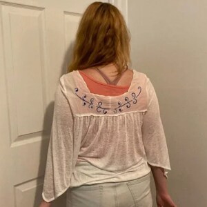 May include: White sheer blouse with a blue floral embroidery design on the back. The blouse has a scoop neck and 3/4 sleeves.