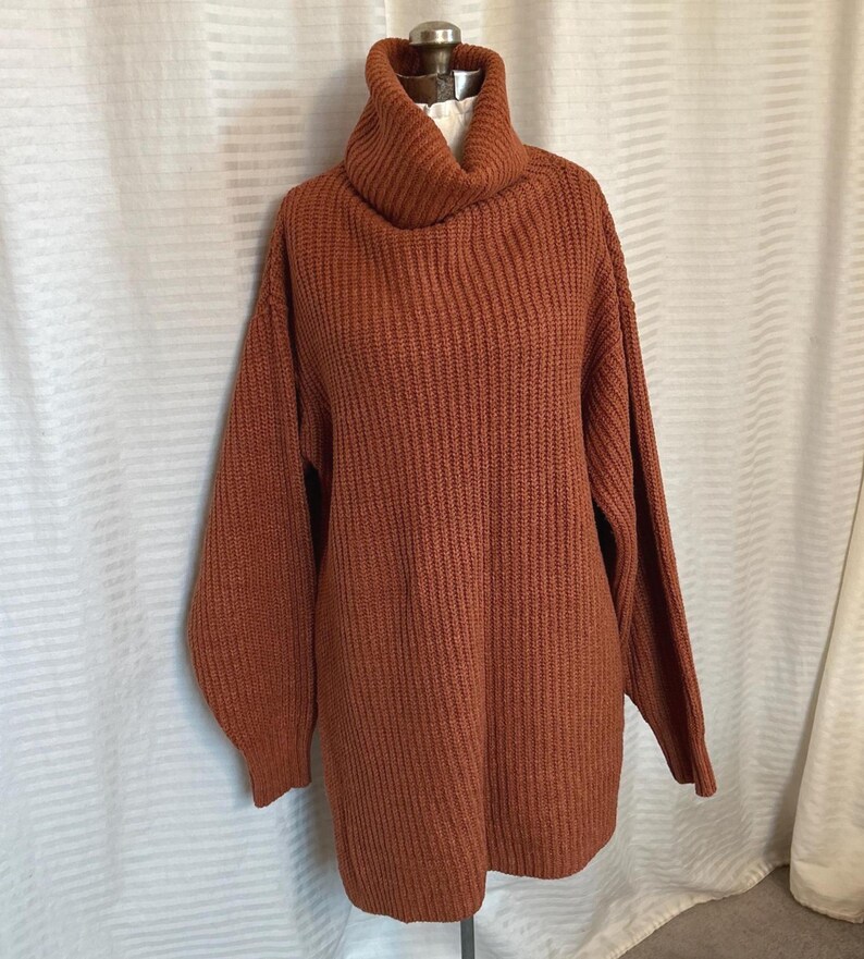 May include: A rust-colored, oversized sweater dress with a ribbed knit texture and a turtleneck collar. The long sleeves have a slight balloon shape. The dress is displayed on a mannequin against a white background.