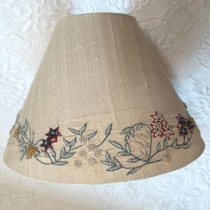 K&ouml;nnte beinhalten: Beigefarbener Lampenschirm mit gesticktem Blumendesign. Der Lampenschirm hat eine konische Form und ist mit einem Rand aus gestickten Blumen in Rot, Gold und Schwarz mit gr&uuml;nen und silbernen Bl&auml;ttern verziert.