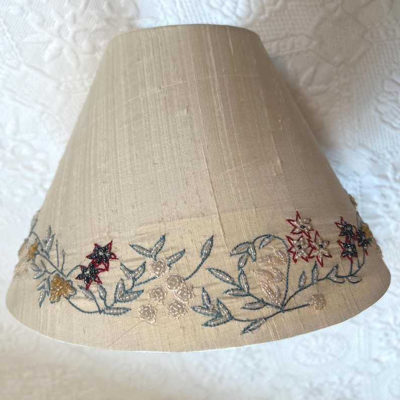 K&ouml;nnte beinhalten: Beigefarbener Lampenschirm mit gesticktem Blumendesign. Der Lampenschirm hat eine konische Form und ist mit einem Rand aus gestickten Blumen in Rot, Gold und Schwarz mit gr&uuml;nen und silbernen Bl&auml;ttern verziert.