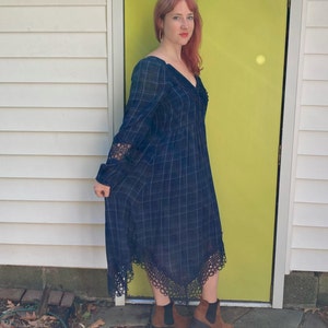 K&ouml;nnte beinhalten: Eine Frau tr&auml;gt ein lang&auml;rmliges, marineblaues Plaid-Kleid mit V-Ausschnitt und Spitzendetails an den &Auml;rmeln und am Saum. Sie tr&auml;gt braune Stiefeletten. Der Hintergrund umfasst eine wei&szlig;e Wand und eine leuchtend gelbe T&uuml;r.