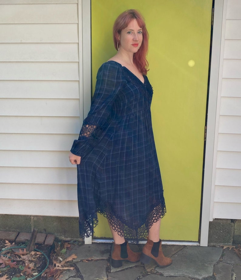 K&ouml;nnte beinhalten: Eine Frau tr&auml;gt ein lang&auml;rmliges, marineblaues Plaid-Kleid mit V-Ausschnitt und Spitzendetails an den &Auml;rmeln und am Saum. Sie tr&auml;gt braune Stiefeletten. Der Hintergrund umfasst eine wei&szlig;e Wand und eine leuchtend gelbe T&uuml;r.