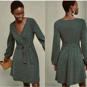 May include: A dark green dress with a white polka dot pattern. The dress has long sleeves, a V-neck, and a belted waist. The dress is knee-length and has a flowing skirt. The model is holding a tan purse.