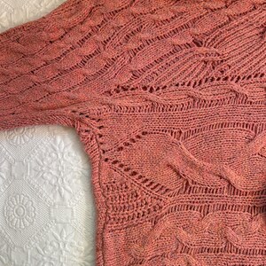 Puede incluir: Un su&eacute;ter de punto color coral con un dise&ntilde;o texturizado. El su&eacute;ter presenta una variedad de patrones de punto, incluyendo trenzas y calados. Las mangas son largas y el cuello es ancho. El su&eacute;ter est&aacute; hecho de un material suave, posiblemente una mezcla de lana.