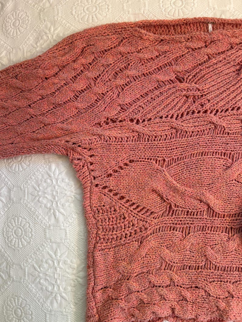 Puede incluir: Un su&eacute;ter de punto color coral con un dise&ntilde;o texturizado. El su&eacute;ter presenta una variedad de patrones de punto, incluyendo trenzas y calados. Las mangas son largas y el cuello es ancho. El su&eacute;ter est&aacute; hecho de un material suave, posiblemente una mezcla de lana.
