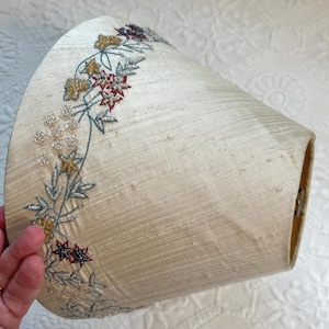 K&ouml;nnte beinhalten: Cremefarbener Lampenschirm mit gesticktem Blumendesign. Die Stickerei weist goldene, rote und blaue Akzente auf. Der Lampenschirm hat eine leicht strukturierte Oberfl&auml;che und eine konische Form, geeignet f&uuml;r eine Tisch- oder Stehlampe.