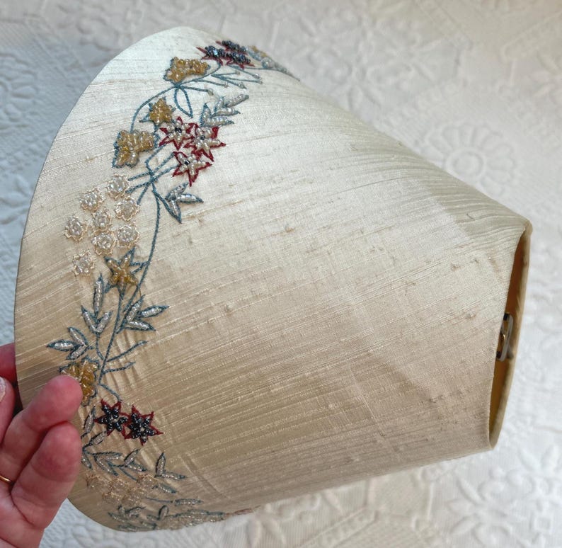 K&ouml;nnte beinhalten: Cremefarbener Lampenschirm mit gesticktem Blumendesign. Die Stickerei weist goldene, rote und blaue Akzente auf. Der Lampenschirm hat eine leicht strukturierte Oberfl&auml;che und eine konische Form, geeignet f&uuml;r eine Tisch- oder Stehlampe.