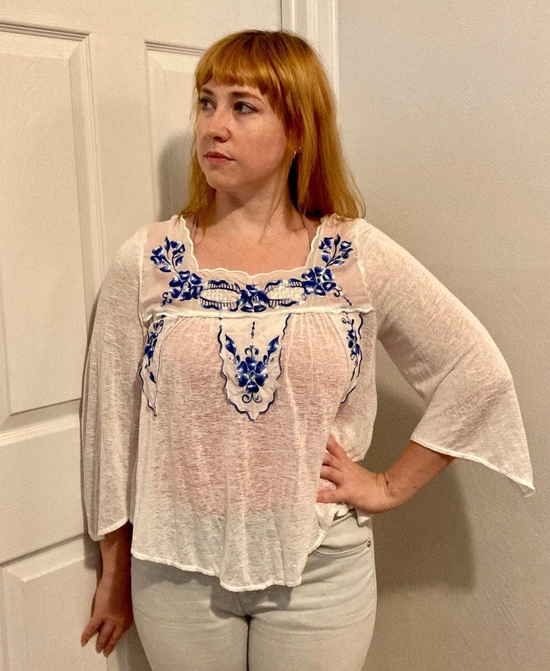 May include: White sheer blouse with blue floral embroidery. The embroidery features a delicate floral pattern with blue and white accents. The blouse has a square neckline and 3/4 sleeves.