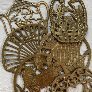 May include: A collection of six overlapping brass trivets featuring various cutout designs including a teapot, a flower basket, a fan, a pineapple, a mountain goat, and an intricate heart-shaped pattern, all with a polished metallic gold finish.