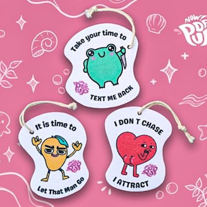 Pode incluir: Três etiquetas caprichosas recortadas com personagens de desenhos animados e frases humorísticas. As etiquetas apresentam um sapo com uma faca, uma manga com boné azul e um coração com pernas. Cada etiqueta tem uma corda para pendurar.