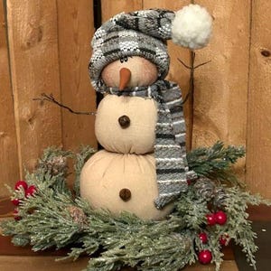 May include: A handmade snowman figurine with a gray and white plaid hat, a gray and white striped scarf, and a brown button nose. The snowman is sitting on a bed of green pine branches with red berries.