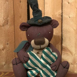 May include: A handmade plush bear with a dark purple body, wearing a dark green hat and holding a green and white striped shamrock. The bear has a tan snout and is sitting down.