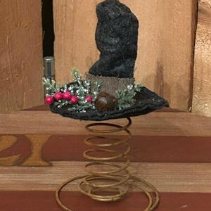 May include: A whimsical, handcrafted decorative item featuring a black felt hat atop a rusty metal spring. The hat is adorned with faux greenery, red berries, and a small acorn. The base is a spiral spring, adding a unique touch.
