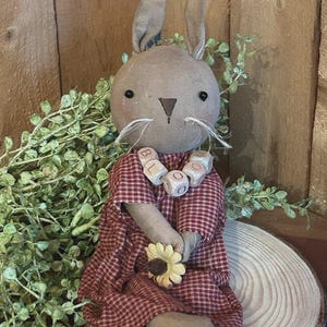 May include: A handmade fabric bunny doll with a brown head, long ears, and a red and white checkered dress. The doll holds a yellow flower and wears a wooden bead necklace spelling "BLOOM". The bunny is seated on a wooden slice, with greenery around it.