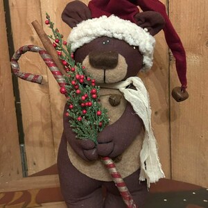 May include: A plush brown bear wearing a burgundy Santa hat with a white trim and a cream-colored scarf. The bear holds a candy cane and a sprig of greenery with red berries, perfect for holiday decor.