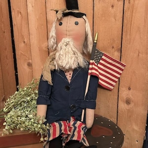 May include: A handmade rag doll dressed in a blue jacket, red and white striped pants, and a black top hat. The doll is holding a small American flag on a stick. The doll is sitting on a wooden stool with a red heart painted on it.