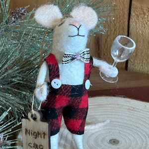 May include: White felt mouse ornament wearing red and black plaid overalls, a bow tie, and holding a small glass of wine. The ornament has a wooden tag that says "Night cap".