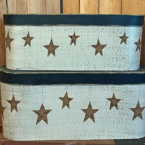 May include: Two oval-shaped, distressed white boxes with navy blue trim. Each box is decorated with brown star cutouts hanging from thin lines. The boxes are stacked on top of each other, creating a rustic, country aesthetic.