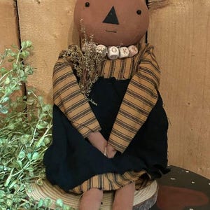 May include: A handmade pumpkin doll with a brown head, black triangle nose, and small eyes. The doll wears a black dress over a striped shirt and has a beaded necklace that spells "OK". The doll is sitting on a wooden surface.