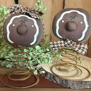 May include: Two brown gingerbread-style figures with painted faces and white icing on a spring base. The figures are on a wooden slice.