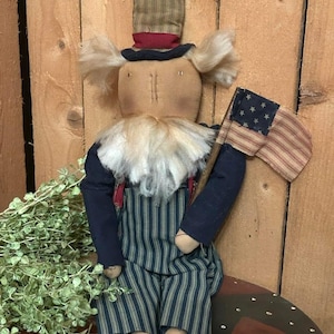 May include: A patriotic doll with a top hat and an American flag. The doll has a tan face, a white beard, and is wearing a blue jacket, striped pants, and red suspenders. The doll is sitting on a decorative wooden surface.