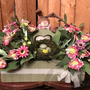 May include: An Easter-themed centerpiece featuring a brown bunny figure peeking over a green box. The box is filled with pink and yellow flowers, green leaves, and a nest with eggs. A white ribbon adds a decorative touch.