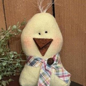 May include: A handmade, plush yellow chick with a brown beak and small black eyes. The chick has rosy cheeks and a plaid scarf with a small wooden button. White wisps of material sprout from the top of its head. A decorative Easter or spring item.