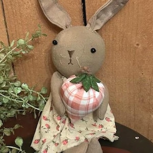 May include: A handmade stuffed bunny doll with a tan body, long ears, and black button eyes. The bunny is holding a plaid strawberry and wearing a floral dress. The doll is approximately 10 inches tall.
