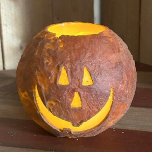 May include: A round, brown Halloween pumpkin with a carved face and a glowing orange interior. The pumpkin has triangular eyes and a curved mouth, creating a smiling expression. The surface has a textured, aged appearance.