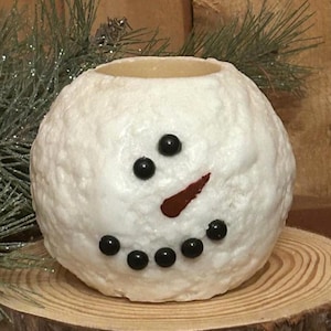 May include: A round, white snowman candle holder with black dot eyes and mouth, and a red carrot nose. The candle holder is sitting on a wooden slice, with green pine needles in the background.