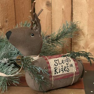 May include: A brown fabric reindeer Christmas decoration with twig antlers, a black nose, and a small bell. The reindeer has a burlap saddle with the words "Sleigh Rides" in black. Green pine branches are attached to the reindeer.