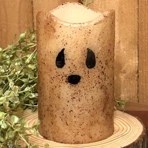 May include: A tan-colored, cylindrical candle with a distressed finish and a black, cartoonish face. The candle sits on a wooden slice, with green foliage in the background. The candle's design evokes a Halloween or fall theme.