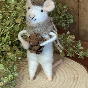 May include: A handcrafted white felt mouse figurine wearing a brown scarf and a small hat. The mouse is holding a brown pinecone. The figurine is standing on a wooden slice, with greenery in the background.