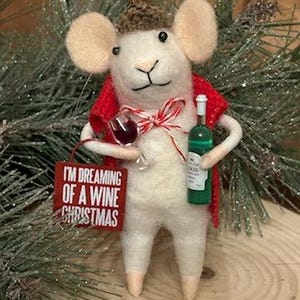 May include: A whimsical felt mouse figurine wearing a red jacket and a small hat. The mouse holds a wine glass and a green bottle, and a sign that reads "I'M DREAMING OF A WINE CHRISTMAS". The figurine is set against a backdrop of evergreen branches.