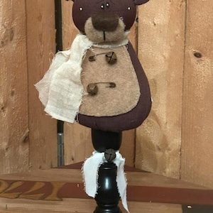 May include: A handcrafted bear figurine with a dark brown head and ears, a tan muzzle, and a cream-colored scarf. The bear has a tan vest with two decorative pins and is mounted on a black wooden stand. The bear is approximately 10 inches tall.