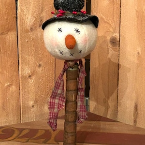 May include: A handmade snowman ornament with a black top hat, red berries, and a red and white plaid scarf. The snowman is made of felt and has a wooden dowel for a body.