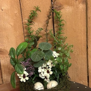 May include: A faux bird's nest with white flowers, green leaves, and two speckled eggs. The nest is made of twigs and is sitting on a wooden surface.