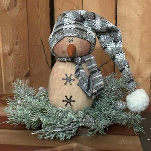 May include: A handcrafted snowman decoration with a gray and white knit hat and scarf. The snowman has a carrot nose and snowflake accents. It is surrounded by frosted greenery and pinecones, creating a festive winter scene.